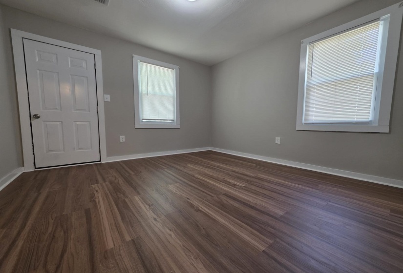 Interior room featuring wood-finish flooring, two windows with white blinds, a white paneled door with silver hardware, and light gray wall paint with white trim