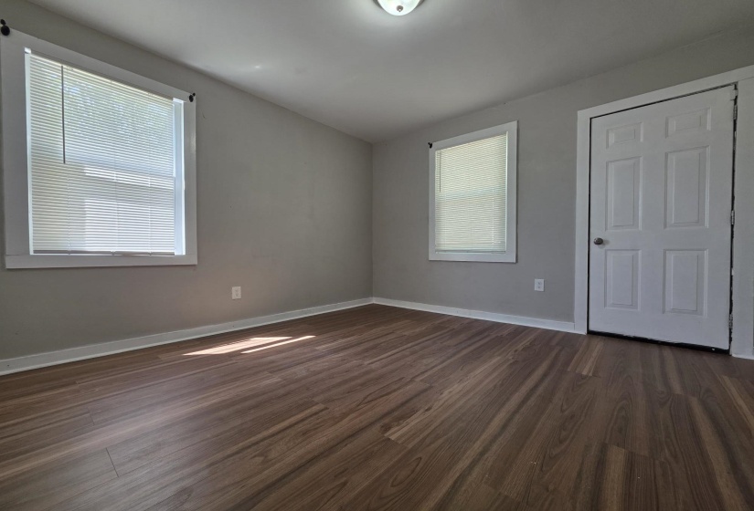 Spacious room featuring wood-finish flooring, light grey wall paint, two windows with blinds, a six-panel door, and white baseboards