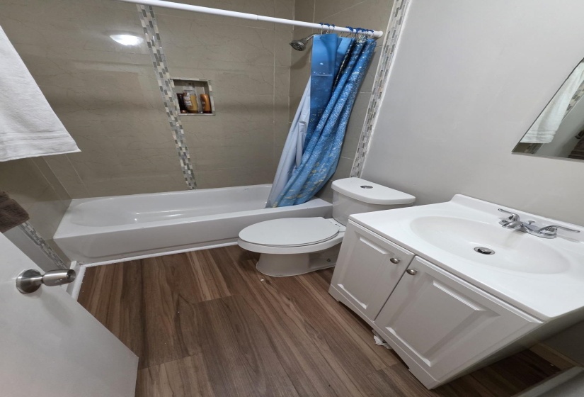 Full bathroom featuring a white bathtub and shower with light-toned tiled surround, an integrated tiled shower niche, wood-finish flooring, and a vanity with a white basin