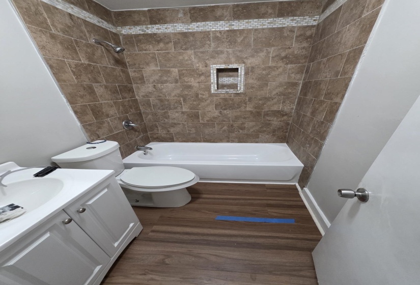 Bathroom featuring wood-finish flooring, a white vanity with under-sink storage, and a shower-tub combination with brown tile surround, an accent tile stripe, and an integrated shower niche