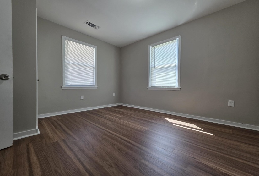 Room featuring wood-finish flooring, two windows with blinds, light gray wall paint, white trim, and a ceiling vent