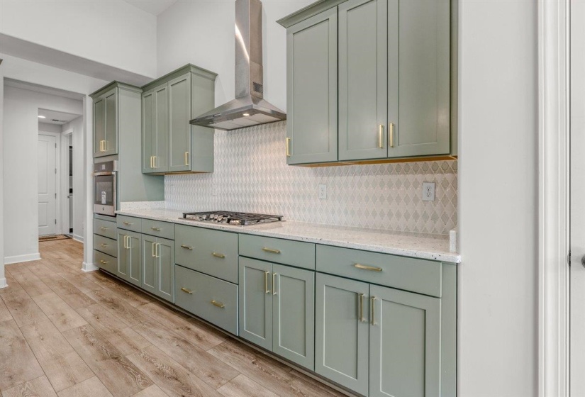 Kitchen featuring sage green cabinetry, a patterned tile backsplash, quartz countertops, a built-in oven, and a gas range with a stainless steel vent hood