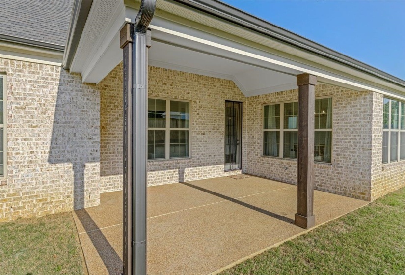 Covered patio featuring a durable aggregate surface, light-colored brick exterior, dark wood-tone support columns, and multiple pane windows