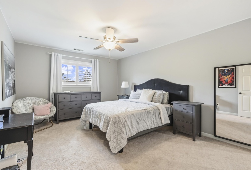 Carpeted bedroom featuring a window with white blinds and drapes, a ceiling fan with integrated lighting, and light gray walls