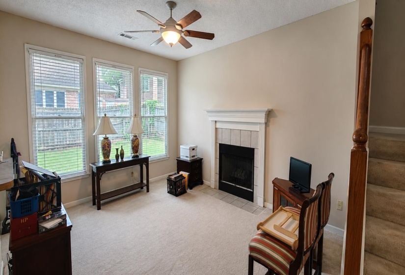 Carpeted living area featuring a fireplace with a tiled surround and mantel, multiple windows, a ceiling fan with integrated lighting, and a wooden staircase with a banister