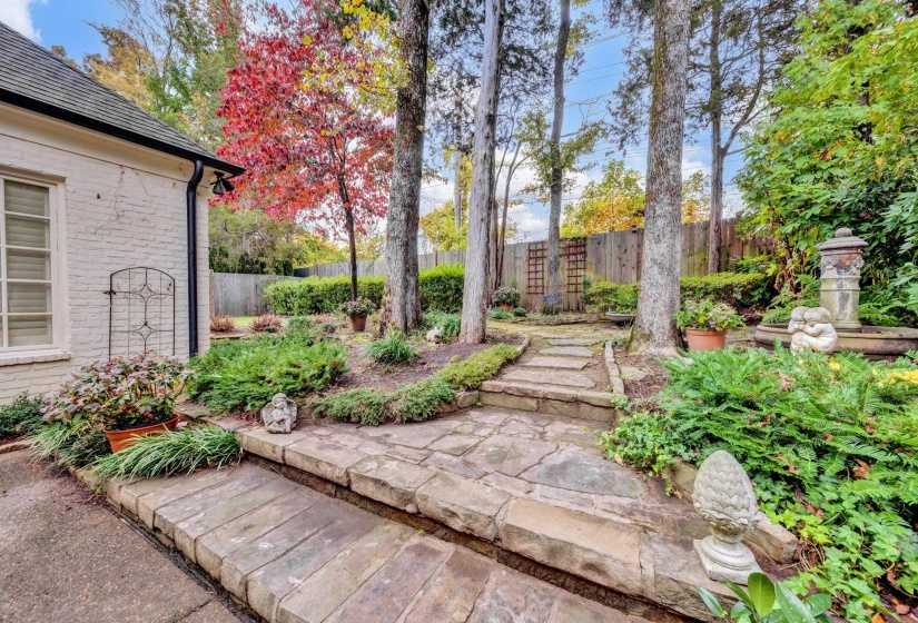 Stone pathway and steps lead through a landscaped garden featuring mature trees, diverse plantings, a privacy fence, and a decorative stone fountain.
