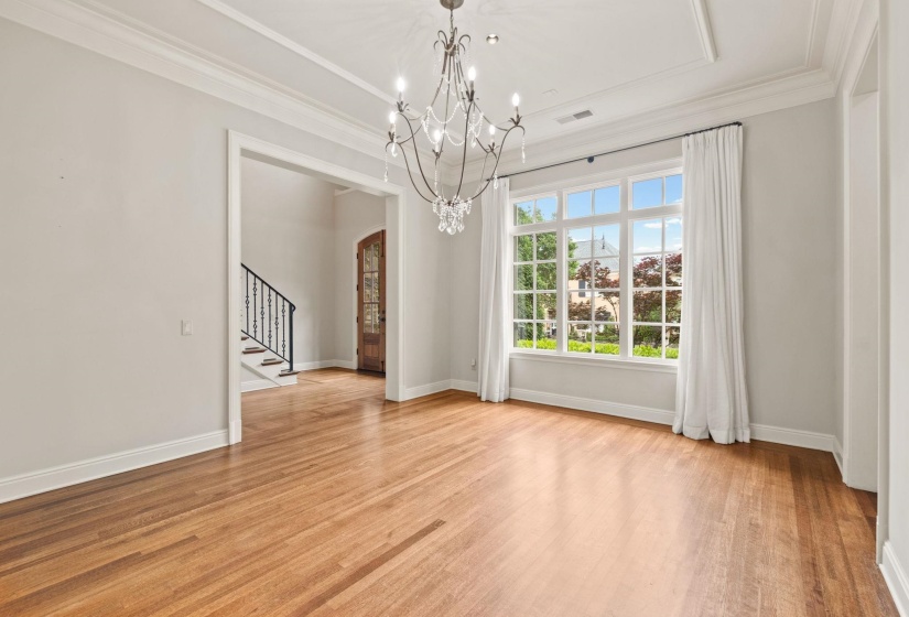 Formal dining room featuring rich hardwood floors, elegant crown molding, sparkling chandelier, and a sun-filled window with serene outdoor views seamlessly connects to the butler’s pantry and flows into the living room for effortless entertaining.
