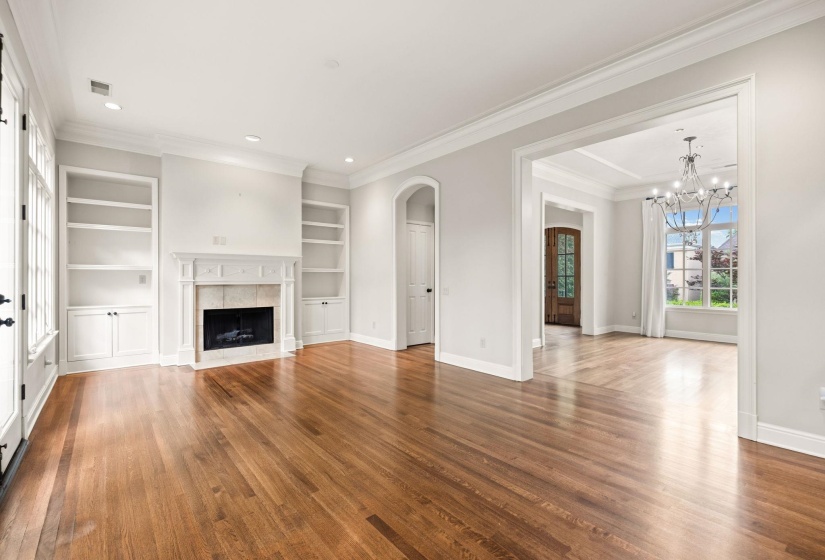 Elegant living room featuring gleaming hardwood floors with walnut inlay, custom built-ins, a classic stone fireplace, and a wall of windows with French doors opening to the back patio, with effortless transition into the home's foyer and dining room.