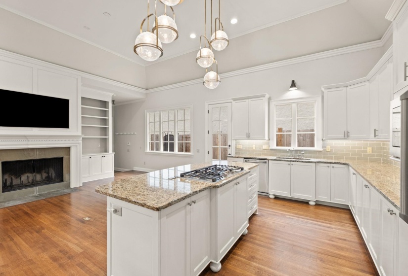 Expansive open-concept kitchen and living area featuring vaulted ceilings, hardwood floors, stainless steel appliances, and a glowing fireplace with detailed mantel. Kitchen sink window overlooks the side yard and adds charm and natural light.