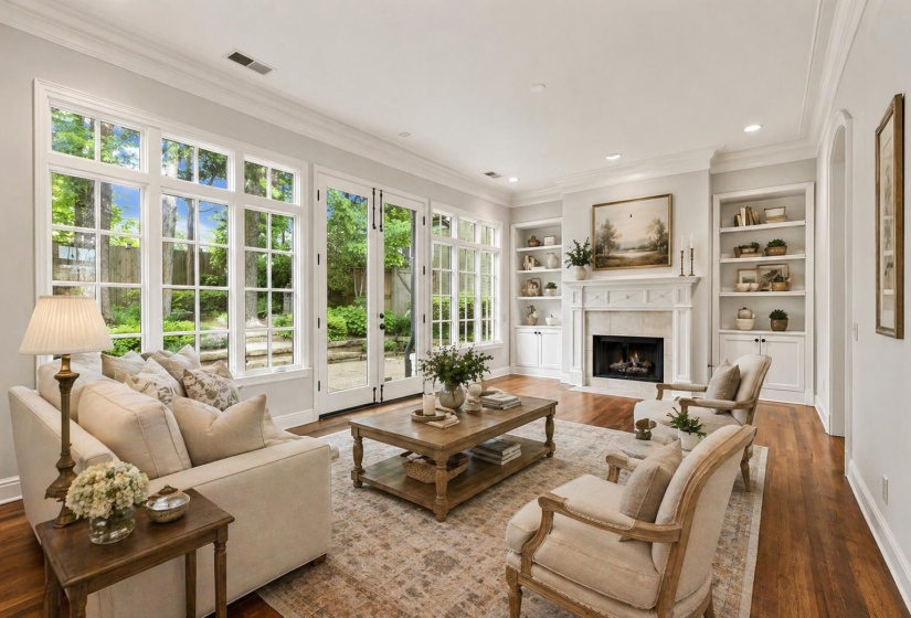 Light-filled living room with expansive paneled windows and French doors overlooking the beautiful garden, a stone fireplace, custom built-ins, hardwood floors, and recessed lighting—ideal for entertaining or quiet evenings at home. Virtually staged.