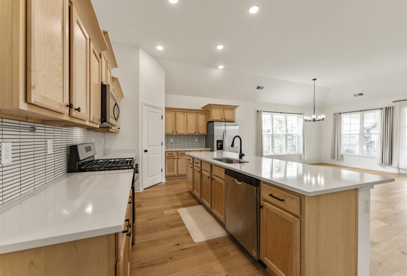 Open-concept kitchen featuring light wood cabinetry, white countertops, a tiled backsplash, stainless steel appliances, and wood-finish flooring