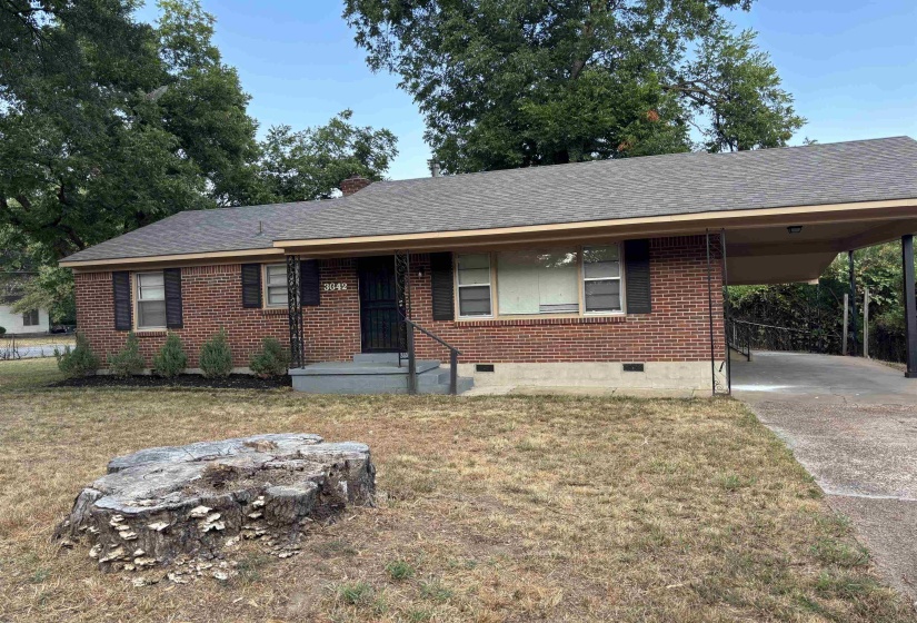 Brick exterior residence featuring a covered carport, dark shingle roof, front entry with security screen door, and foundational plantings