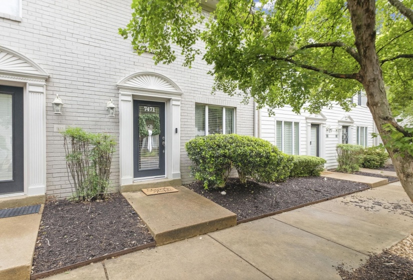 Townhome exterior featuring light brick facade, dark entry door with transom, fluted trim, and mature landscaping