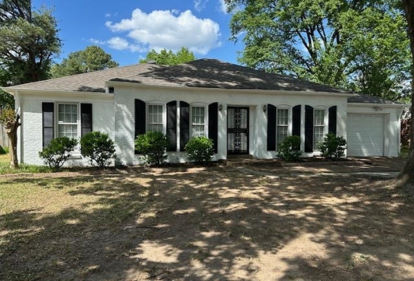 White brick exterior with a dark shingle roof, featuring black window shutters, a decorative security door, and an integrated garage