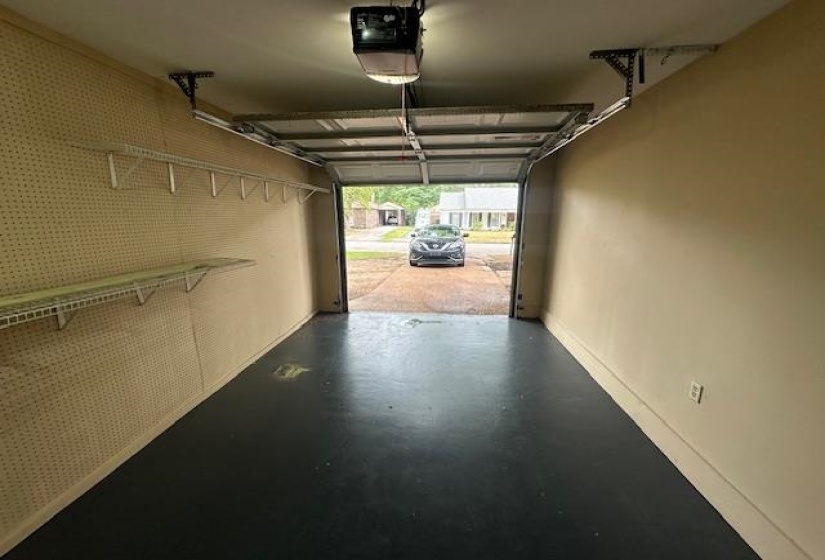 Single-car garage featuring a dark-colored floor, automatic garage door opener, and pegboard walls with wire shelving