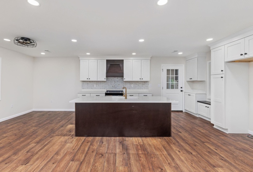 Kitchen featuring custom exhaust hood, backsplash, white cabinetry, an island with sink, and light countertops