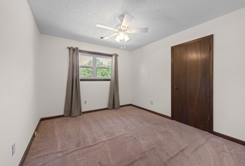 Carpeted room with a ceiling fan, a window with exterior tree views, white walls, and a dark wood-finish door