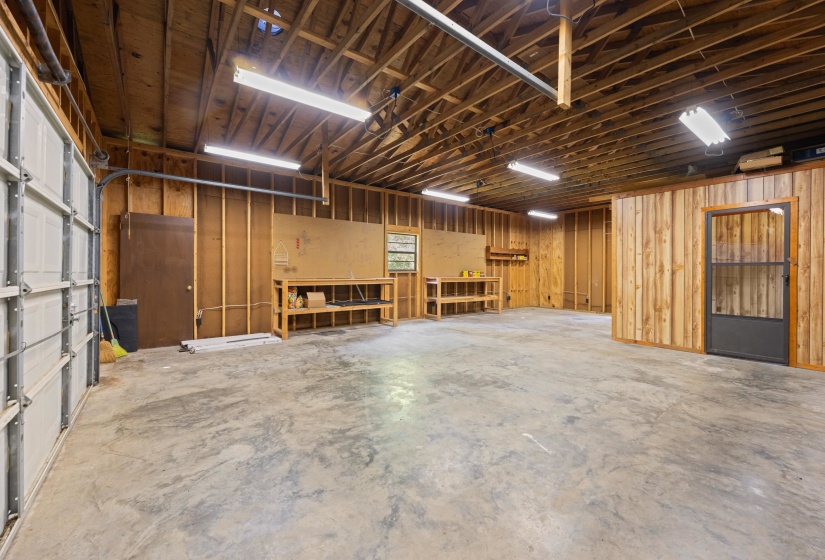 Spacious garage featuring a concrete floor, exposed wood ceiling joists, fluorescent lighting, a roll-up garage door, and a dedicated utility area with built-in shelving