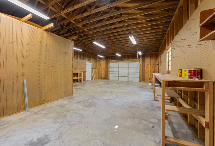 Spacious garage featuring a concrete floor, exposed wood beam ceiling, multiple fluorescent light fixtures, a rolling garage door, and built-in workbench shelving