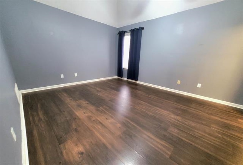 Spacious room featuring wood-finish flooring, a window with dark curtains, light gray walls, white trim, and a vaulted ceiling