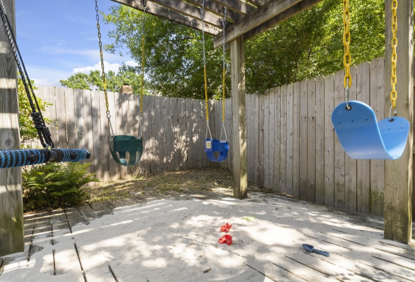 Shaded outdoor play area featuring a wooden pergola structure, multiple swings, a sand-filled ground, and a wooden privacy fence