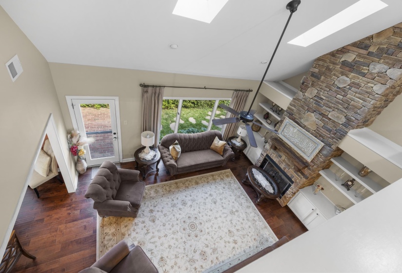 Vaulted ceiling living area featuring a stone-faced fireplace, built-in shelving, wood-finish flooring, and dual skylights