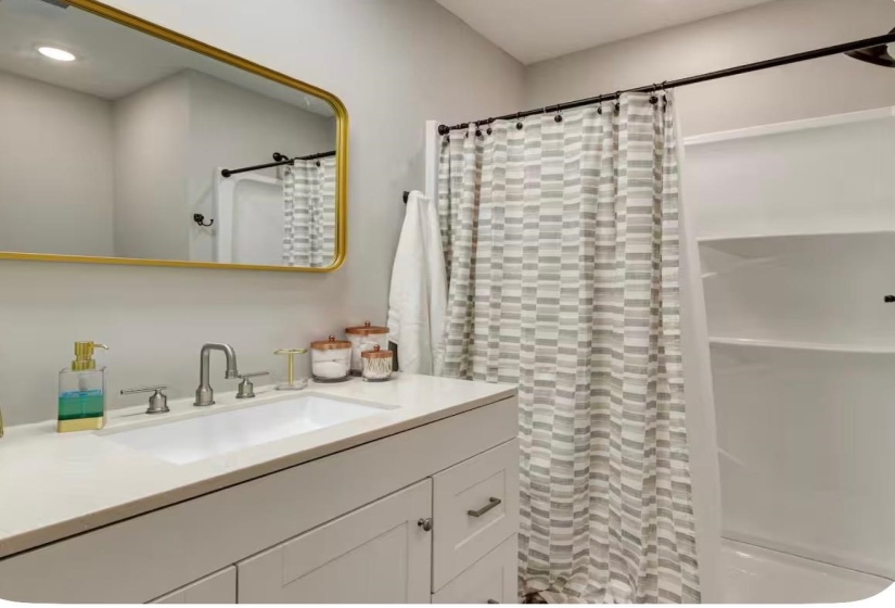 Bathroom featuring a rectangular gold-framed mirror, an integrated sink with a brushed nickel faucet, a white vanity with paneled doors and drawer, and a shower-tub combination with built-in shelving