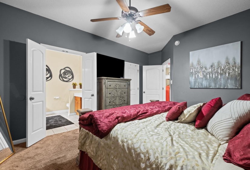 Carpeted bedroom featuring a ceiling fan with light fixture, gray painted walls, and double white paneled doors opening to a tiled area with a light wood vanity