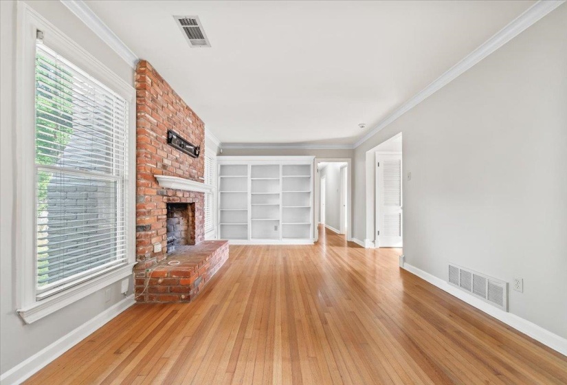 Living area featuring wood-finish flooring, a brick fireplace with hearth, white built-in shelving, white trim, and a window with blinds
