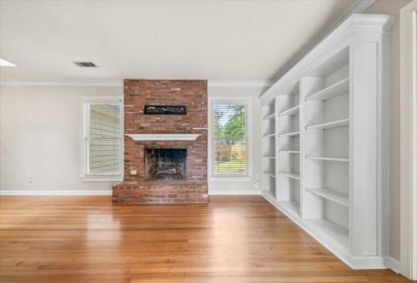 Hardwood flooring throughout featuring a brick fireplace with a white mantel, built-in white shelving, and crown molding