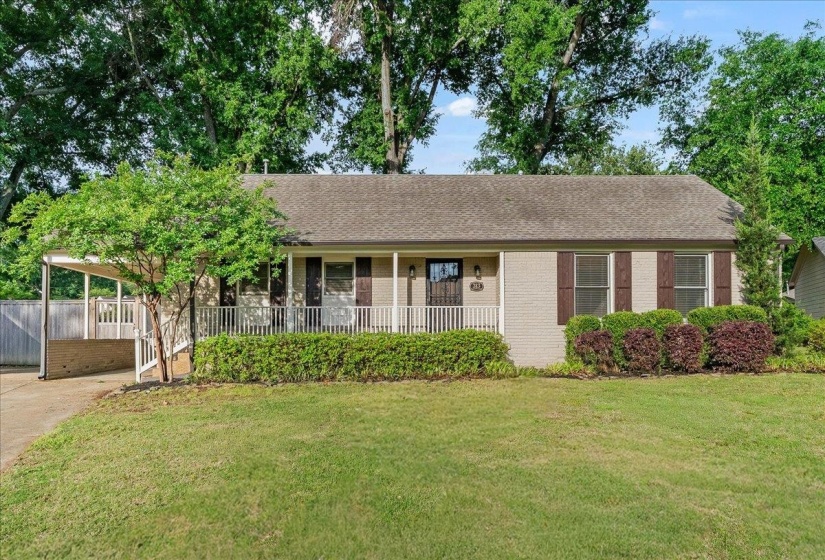 Single-story residence featuring a covered front porch with white railing, light-colored brick exterior, dark window shutters, and a carport
