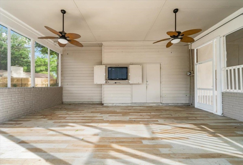 Expansive screened porch featuring painted brick half-walls, wood-finish flooring, two ceiling fans with integrated lighting, and a built-in media cabinet
