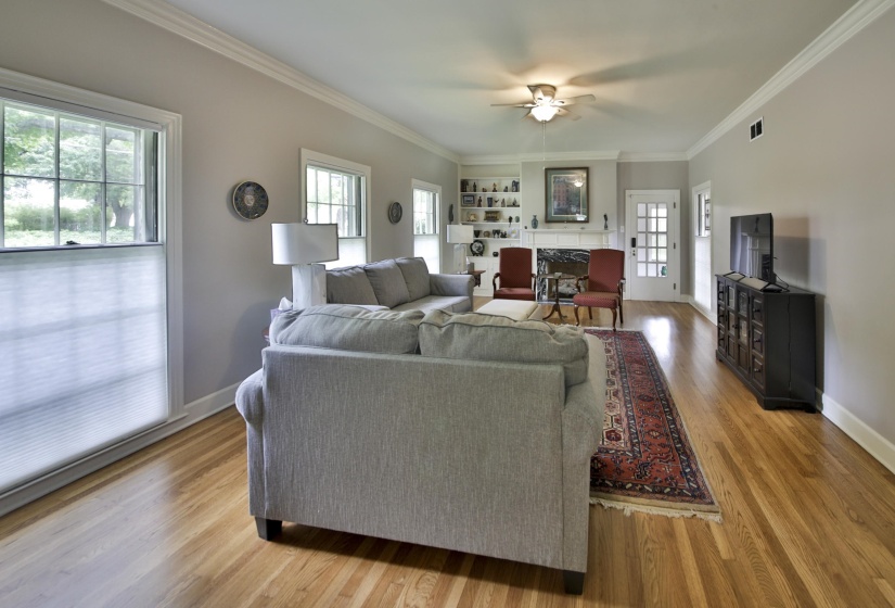 Spacious living area featuring wood-finish flooring, a fireplace with a white mantel, built-in shelving, a ceiling fan, and multiple windows