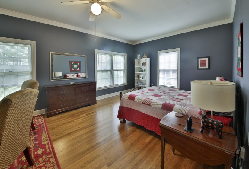 Spacious room featuring hardwood flooring, multiple windows providing ample natural light, crown molding, a ceiling fan, and a built-in shelving unit
