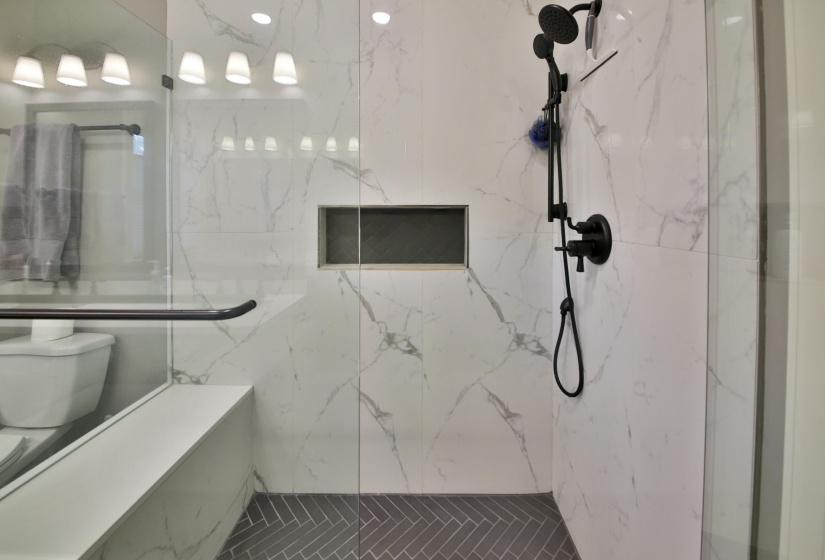 Spacious walk-in shower featuring glass enclosure, white marble-look tile walls, gray herringbone floor tile, built-in bench, and a recessed shower niche