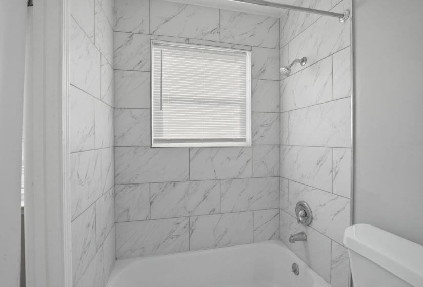 Bathroom featuring white rectangular wall tiles with subtle veining, a built-in bathtub, a window with blinds, and chrome fixtures including a showerhead and tub spout