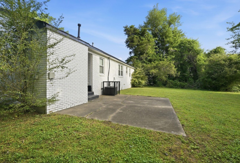Spacious backyard featuring a concrete patio, expansive lawn, white brick exterior, black roofline, and established perimeter trees