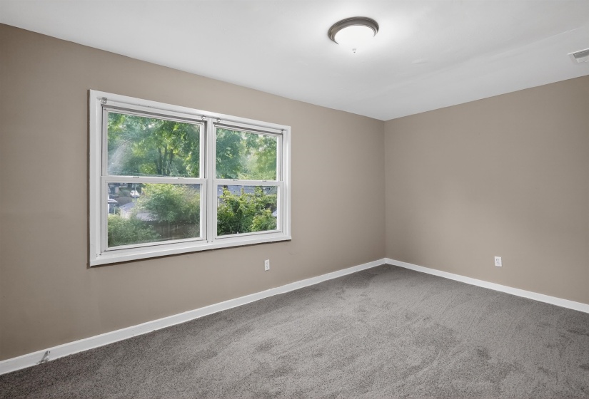 Neutral wall paint, plush carpeting, and white trim define this room, featuring a double window with a wooded backdrop and a flush-mount ceiling light