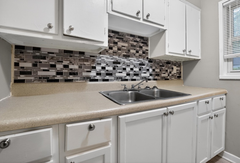 Kitchen featuring white cabinetry, a dual-basin stainless steel sink, a mosaic tile backsplash, light-toned countertops, and a window with blinds