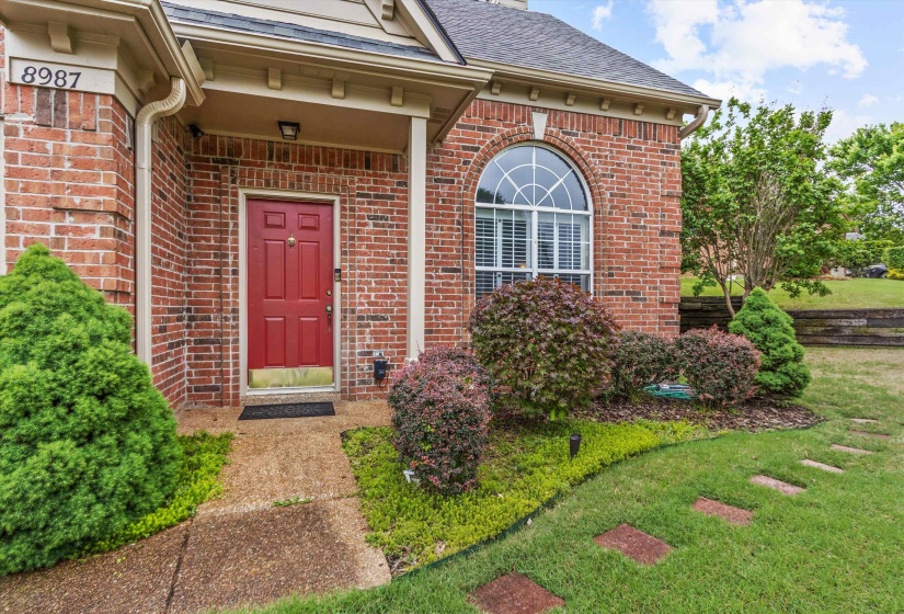 Brick exterior with a red front door, featuring an arched window with white blinds, a paved walkway, and landscaping with mature shrubs and stepping stones