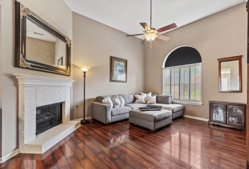 Spacious living area featuring high vaulted ceilings, rich wood-finish flooring, a painted brick fireplace with hearth, an arched window with blinds, and a ceiling fan