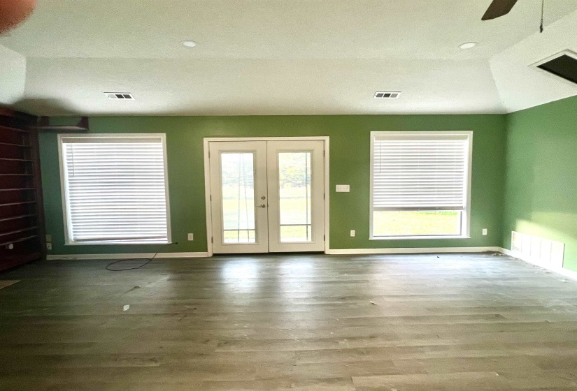 Spacious room with wood-finish flooring, featuring double glass-paneled doors and two large windows with blinds