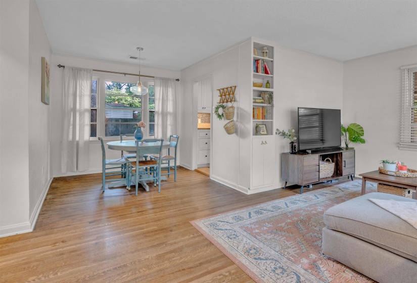 Open-concept living area featuring wood-finish flooring, a dining space with a window, and a built-in shelving unit