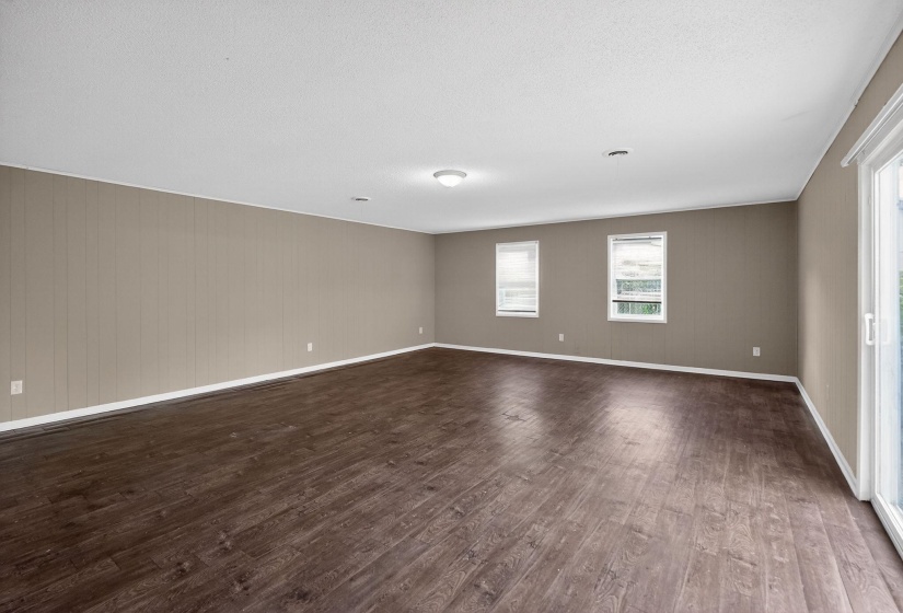 Spacious room featuring wood-finish flooring, two windows with blinds, a sliding glass door, and neutral wall paneling