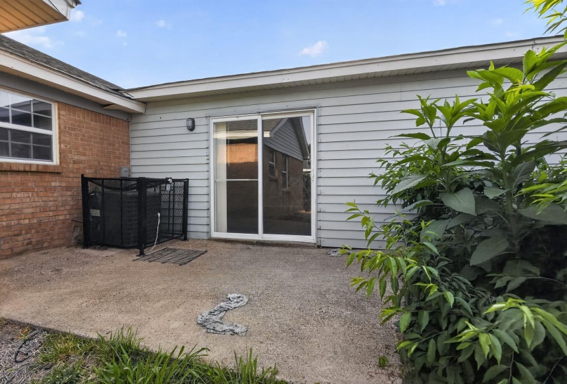 Concrete patio with a sliding glass door, enclosed utility unit, brick exterior accent, and mature perimeter landscaping