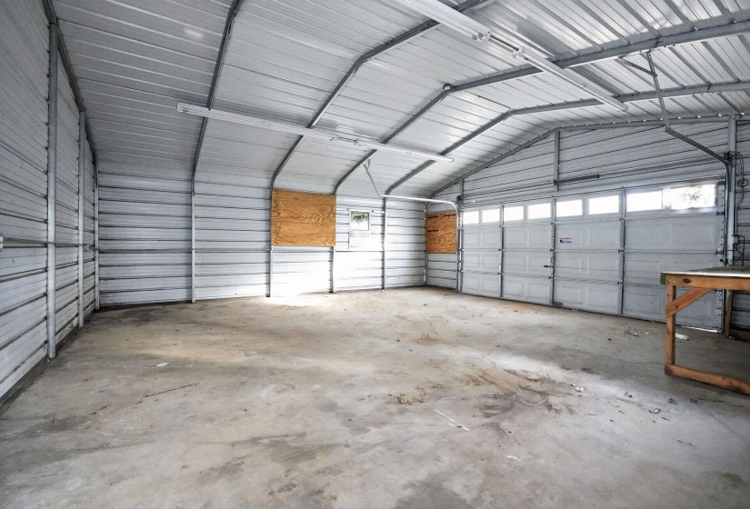 Spacious corrugated metal structure featuring a concrete floor, multiple fluorescent light fixtures, and a large garage door with transom windows