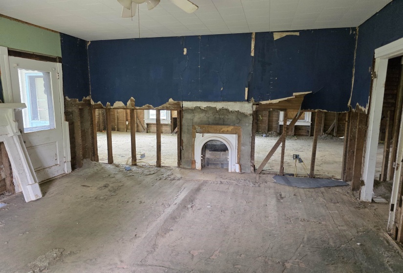 Interior space with exposed wall studs, an original fireplace surround, a partial door frame with trim, a ceiling fan, and exposed subflooring
