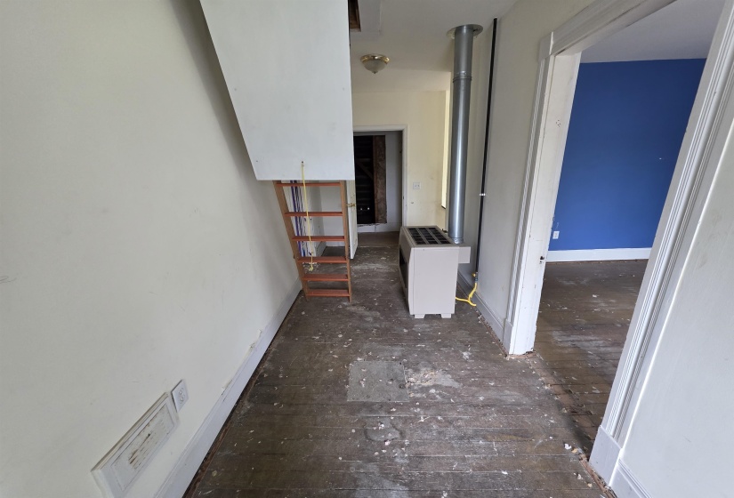 Interior hallway featuring exposed wood subflooring, a built-in furnace unit with vertical venting, and an attic access ladder