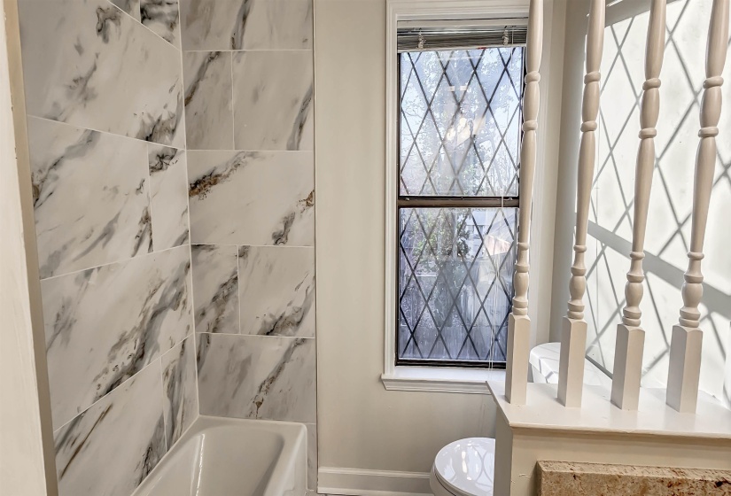 Bathroom featuring a white bathtub with marble-look tile surround, a diamond-patterned window, and white painted balusters
