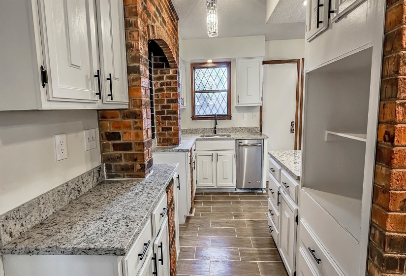 Kitchen featuring white shaker-style cabinetry, light-toned granite countertops, wood-finish tile flooring, a brick arched architectural element, and a stainless steel dishwasher